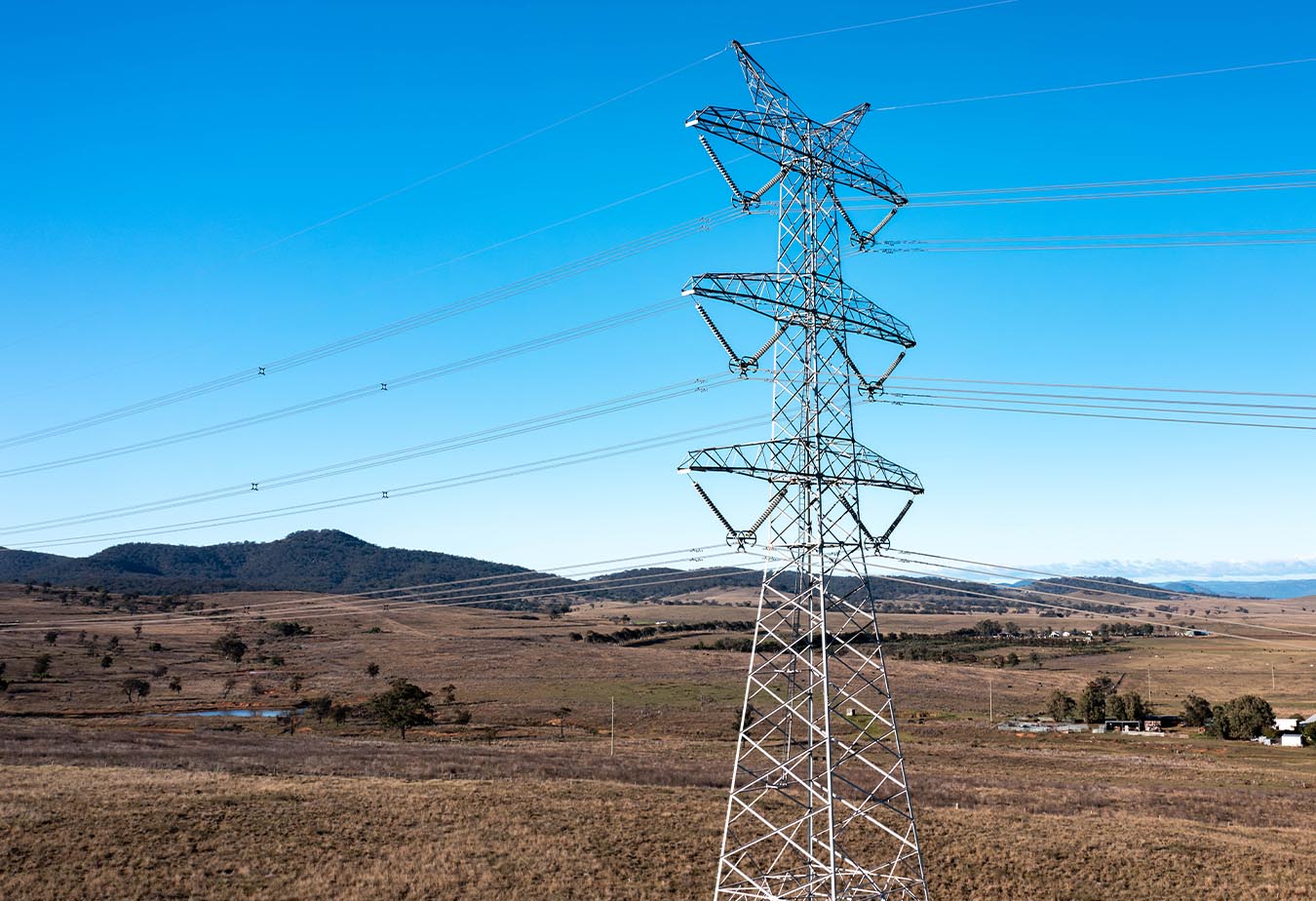 Metal power transmission tower with multiple lines stretching across a rural Australian area with hills and blue sky.