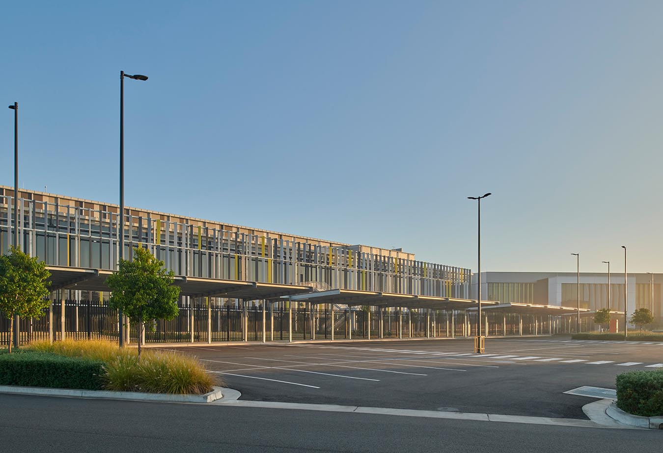 Modern multi-story building with large glass windows, trees and some low bushes, and a paved car-park area in the foreground.