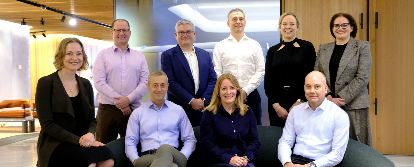 Group photo of our nine Senior Leadership Team members in business attire, posed in an office setting. Some are seated with others standing behind them.