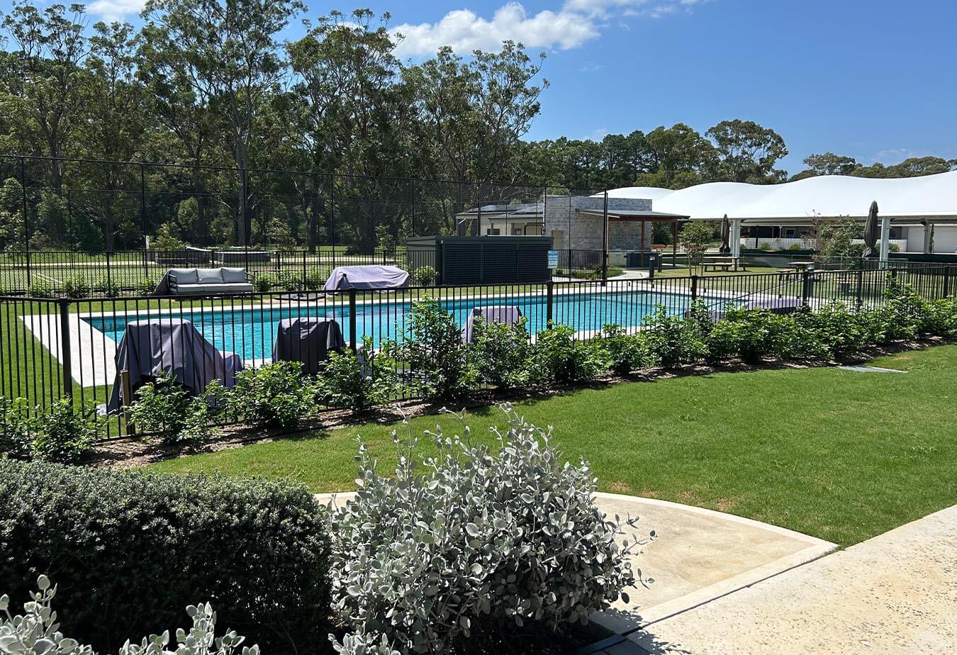 An outdoor swimming pool enclosed by a black metal fence, bordered by trimmed greenery and set against a backdrop of trees and buildings under blue sky.